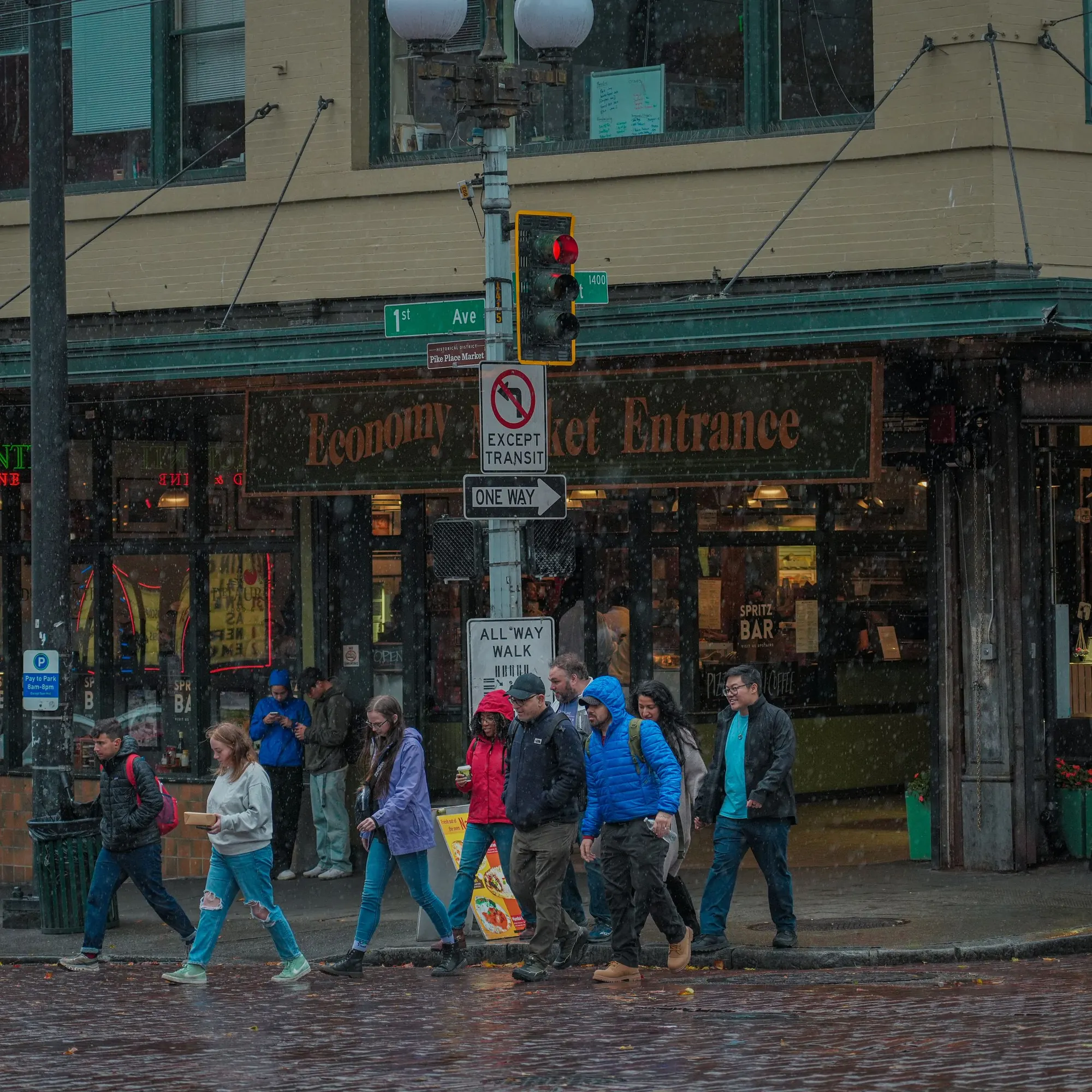 People in rain jackets walk past a market storefront on a wet brick street on a rainy day in Seattle