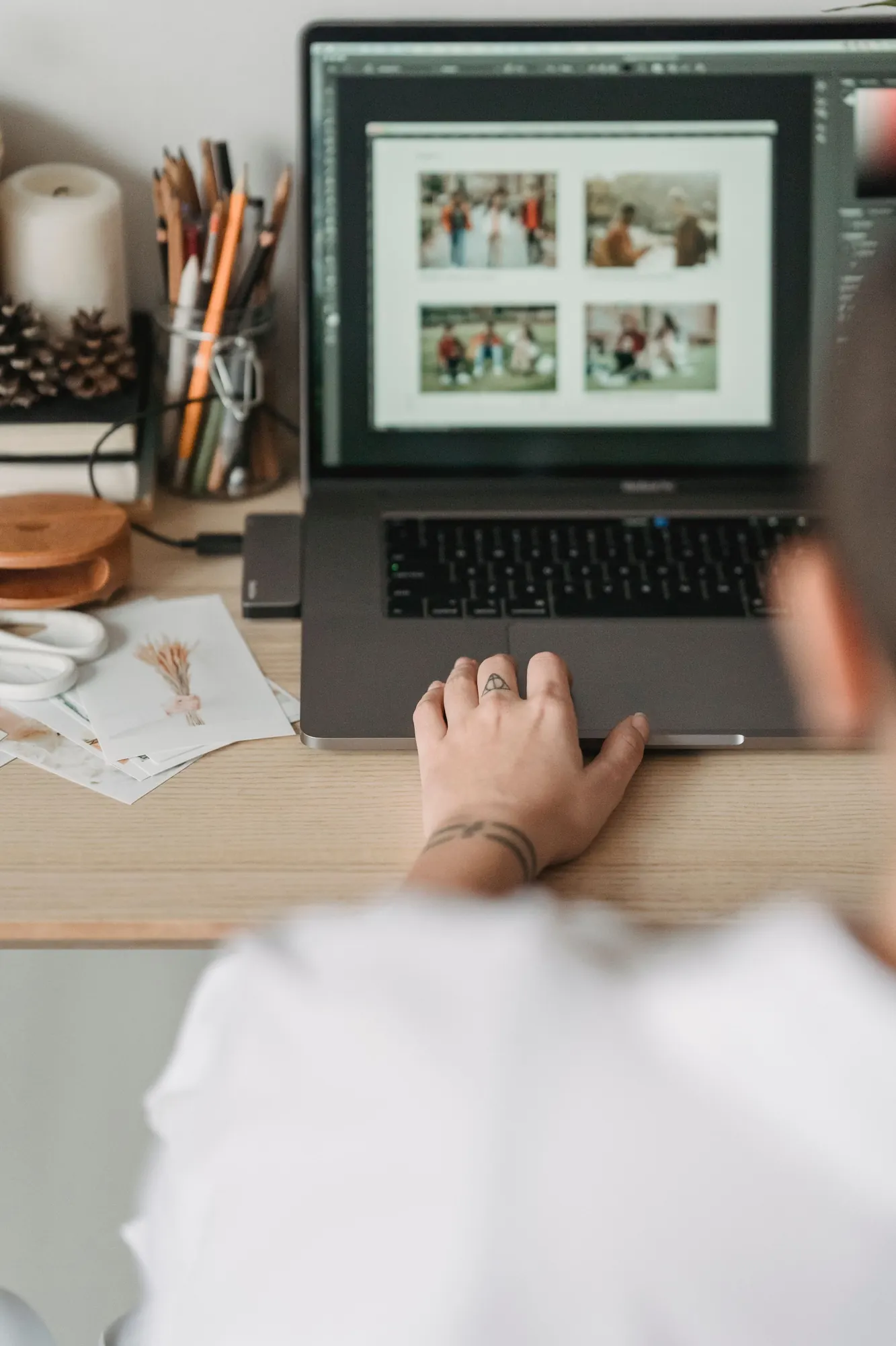 Over-the-shoulder view of a hand on a laptop trackpad, editing a grid of photos, with pencils and prints on a wooden desk