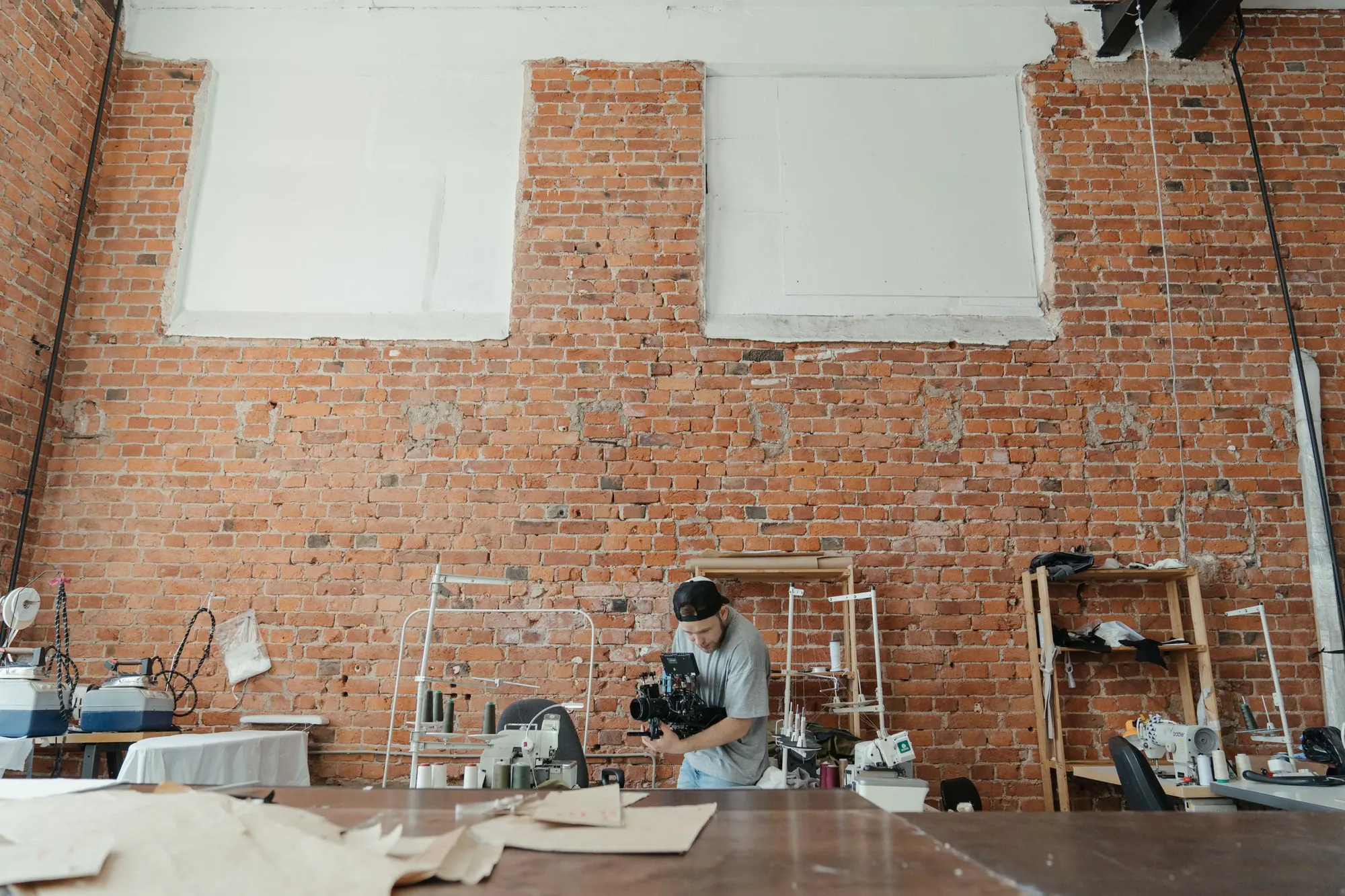 A camera operator films inside a brick-walled sewing workshop with sewing machines, racks and pattern paper on the cutting table
