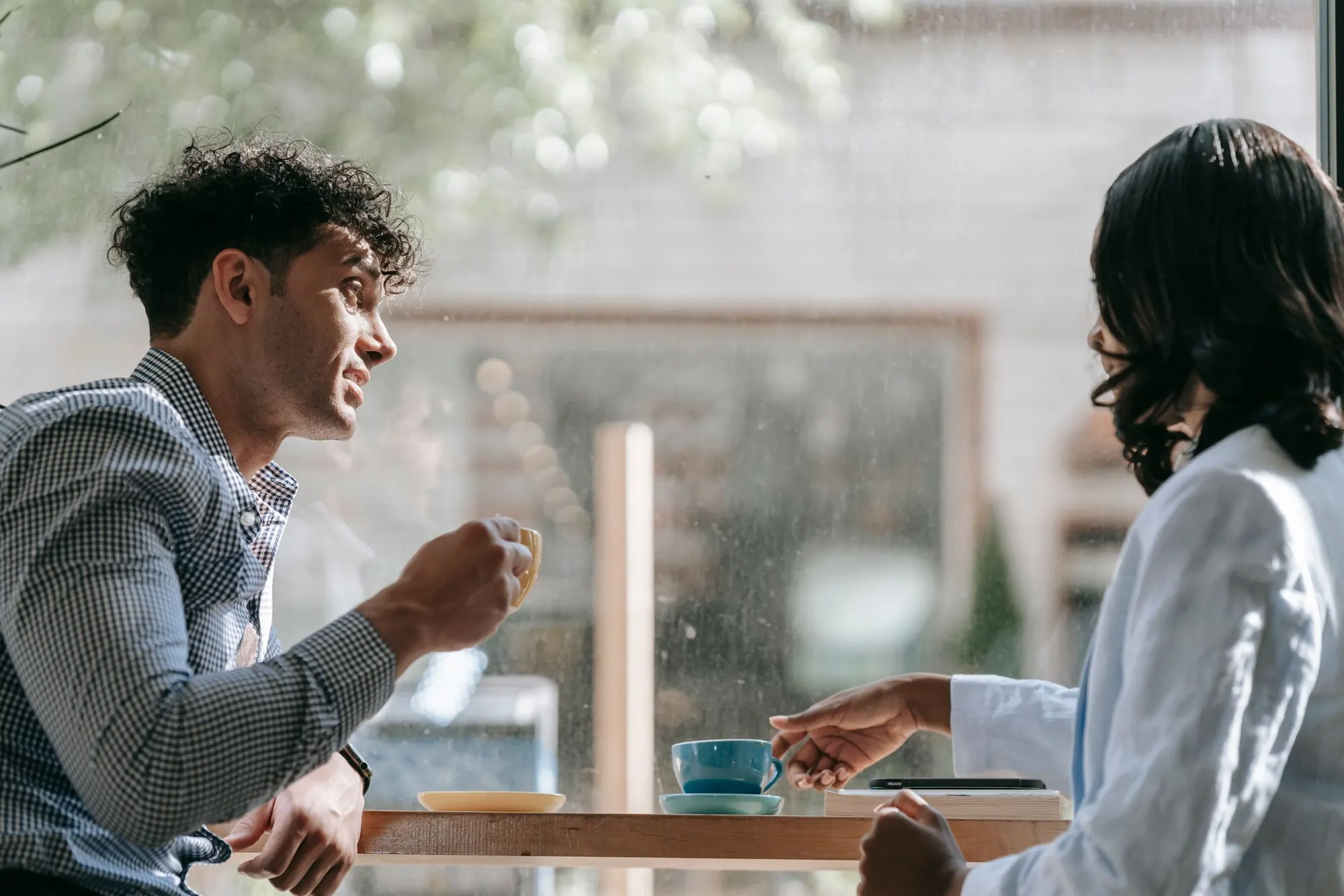 Two people talk over coffee at a window counter, with rain-streaked glass behind them
