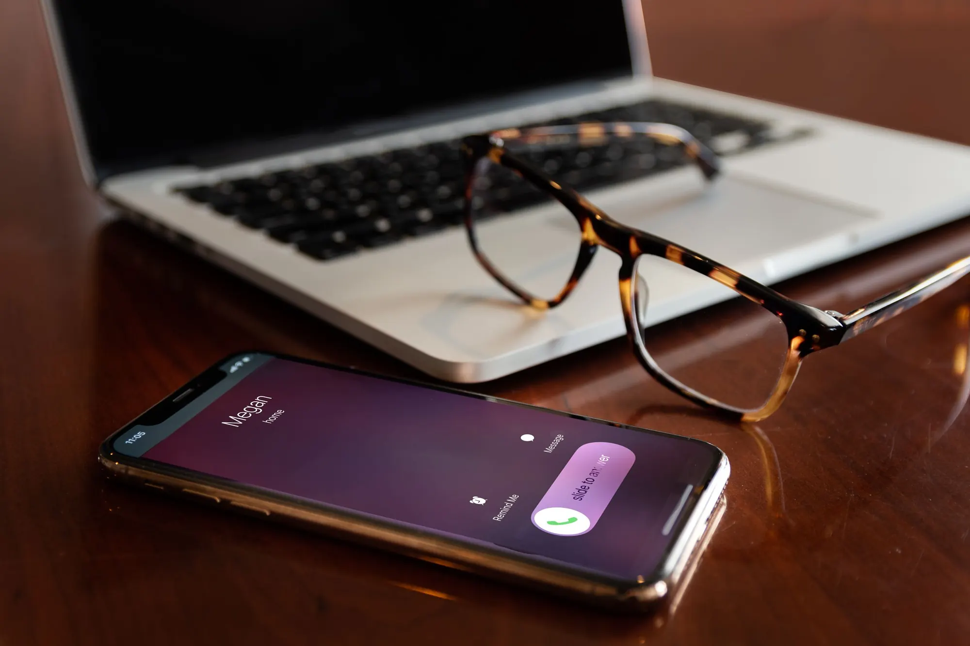 A smartphone shows an incoming call on a wooden desk, next to a laptop and tortoiseshell glasses