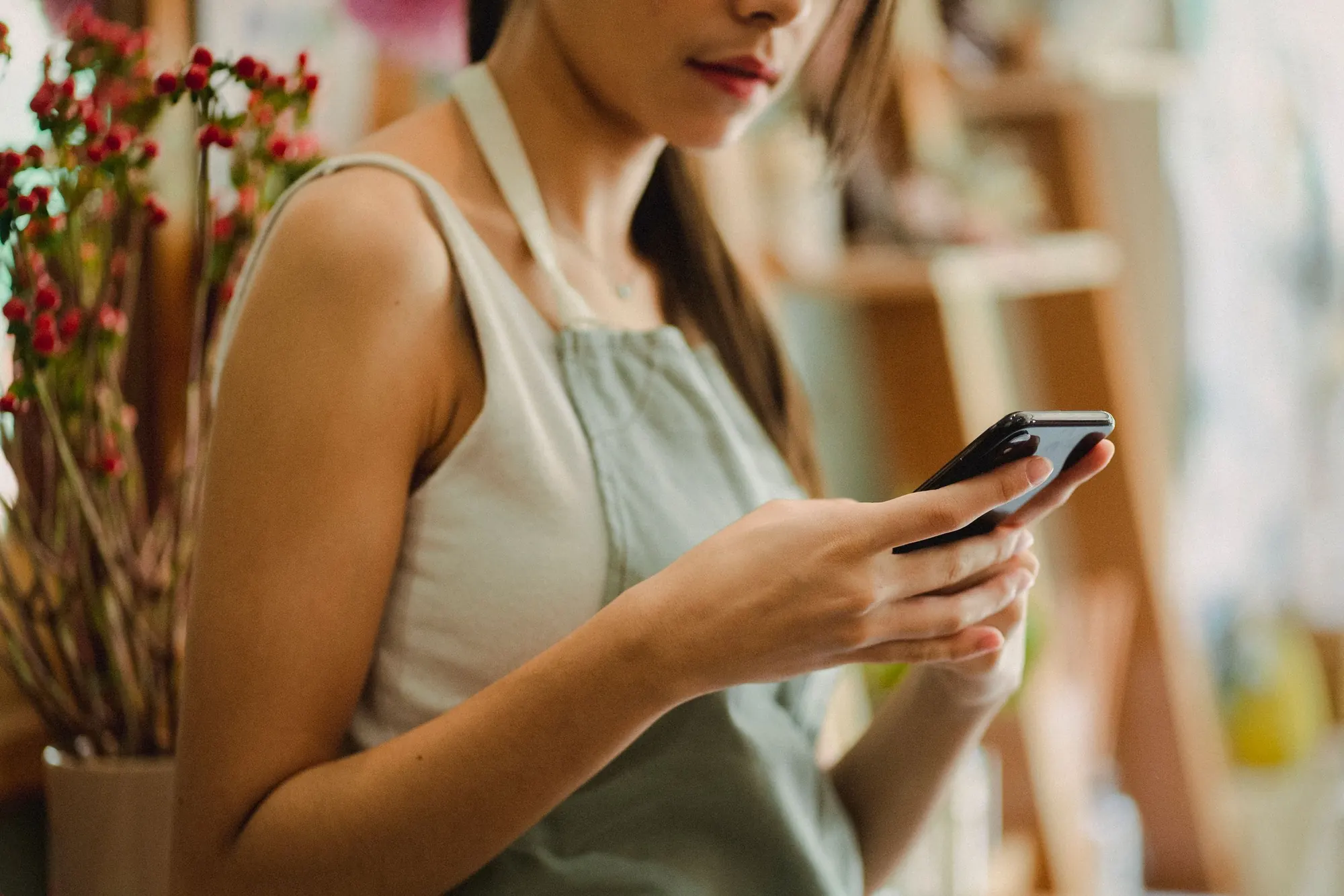 A shop worker in a linen apron reads a message on her phone in a flower shop, her face cropped at the lips