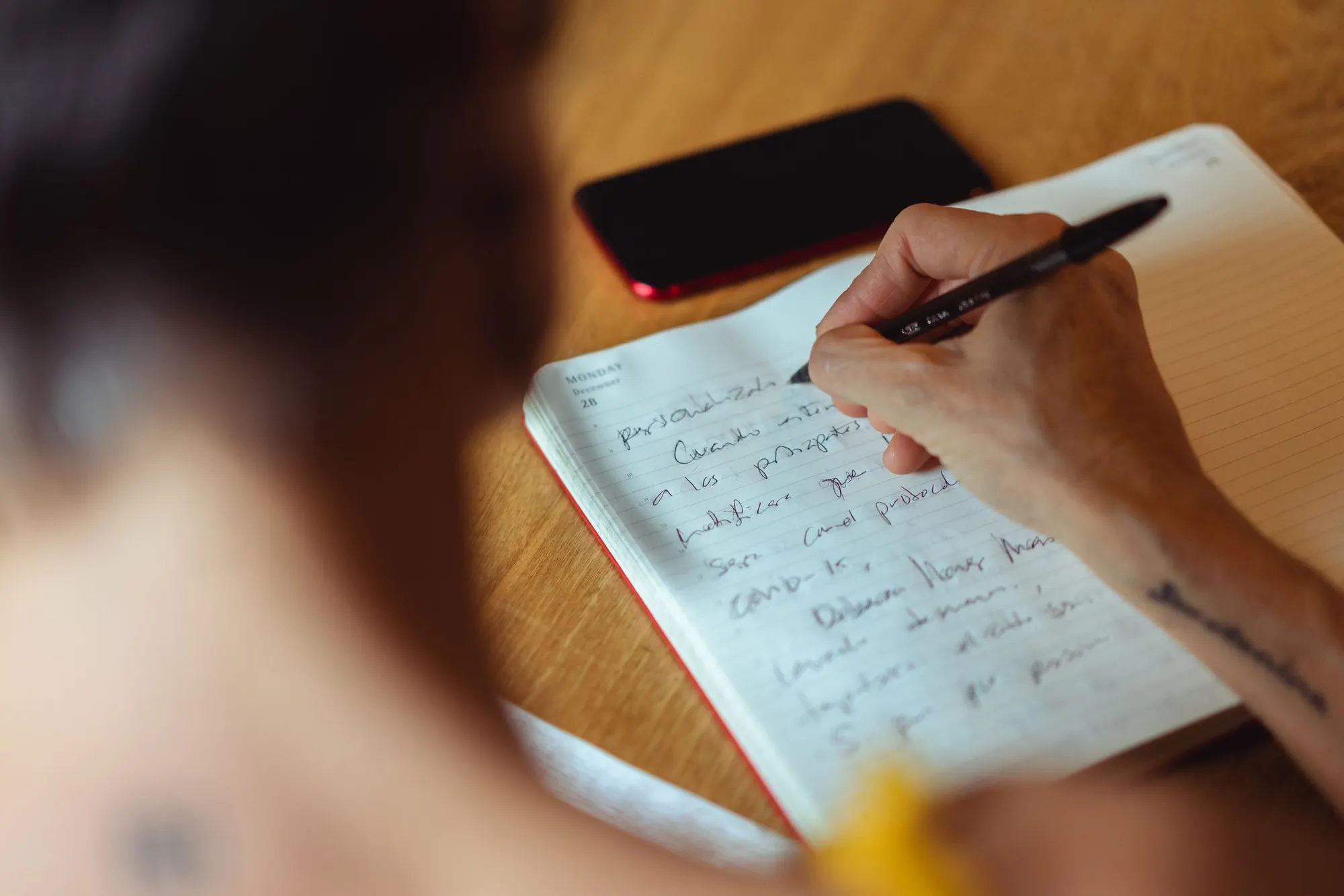 Over-the-shoulder close-up of a hand writing notes in a lined notebook on a wooden table, a phone beside it