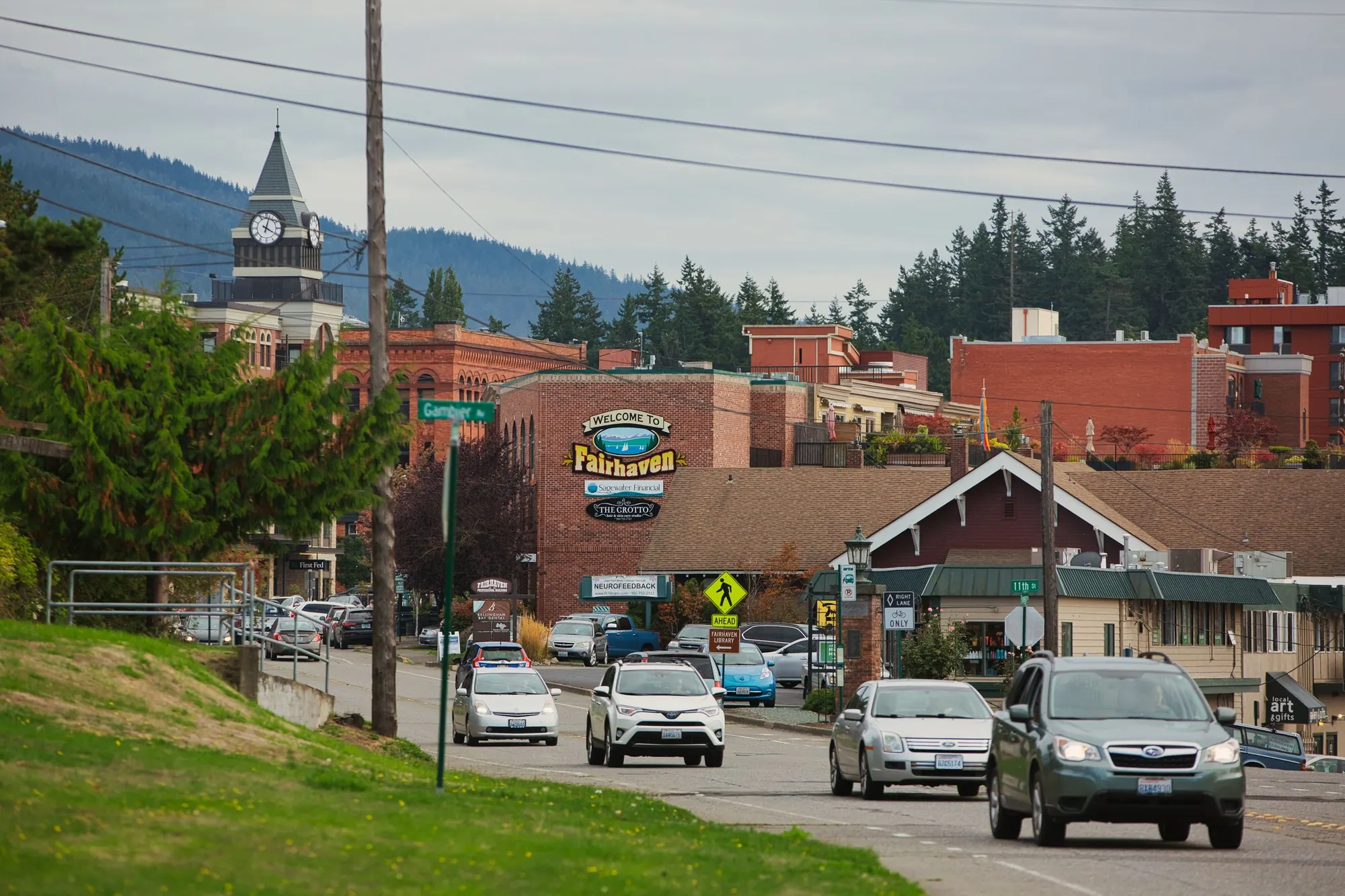A small-town street in the Fairhaven district of Bellingham, WA, with brick buildings, a clock tower, cars and evergreen hills under an overcast sky