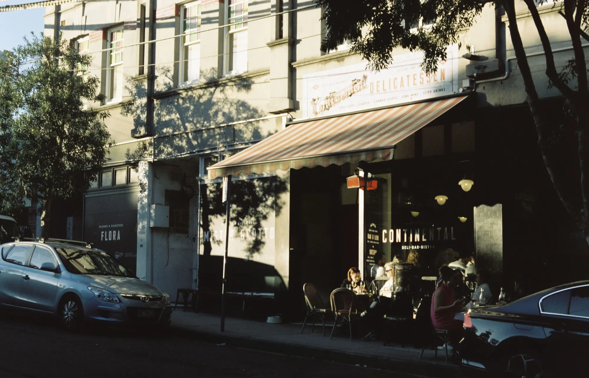 A corner deli with a striped awning and full sidewalk tables in low golden-hour light, shot on film