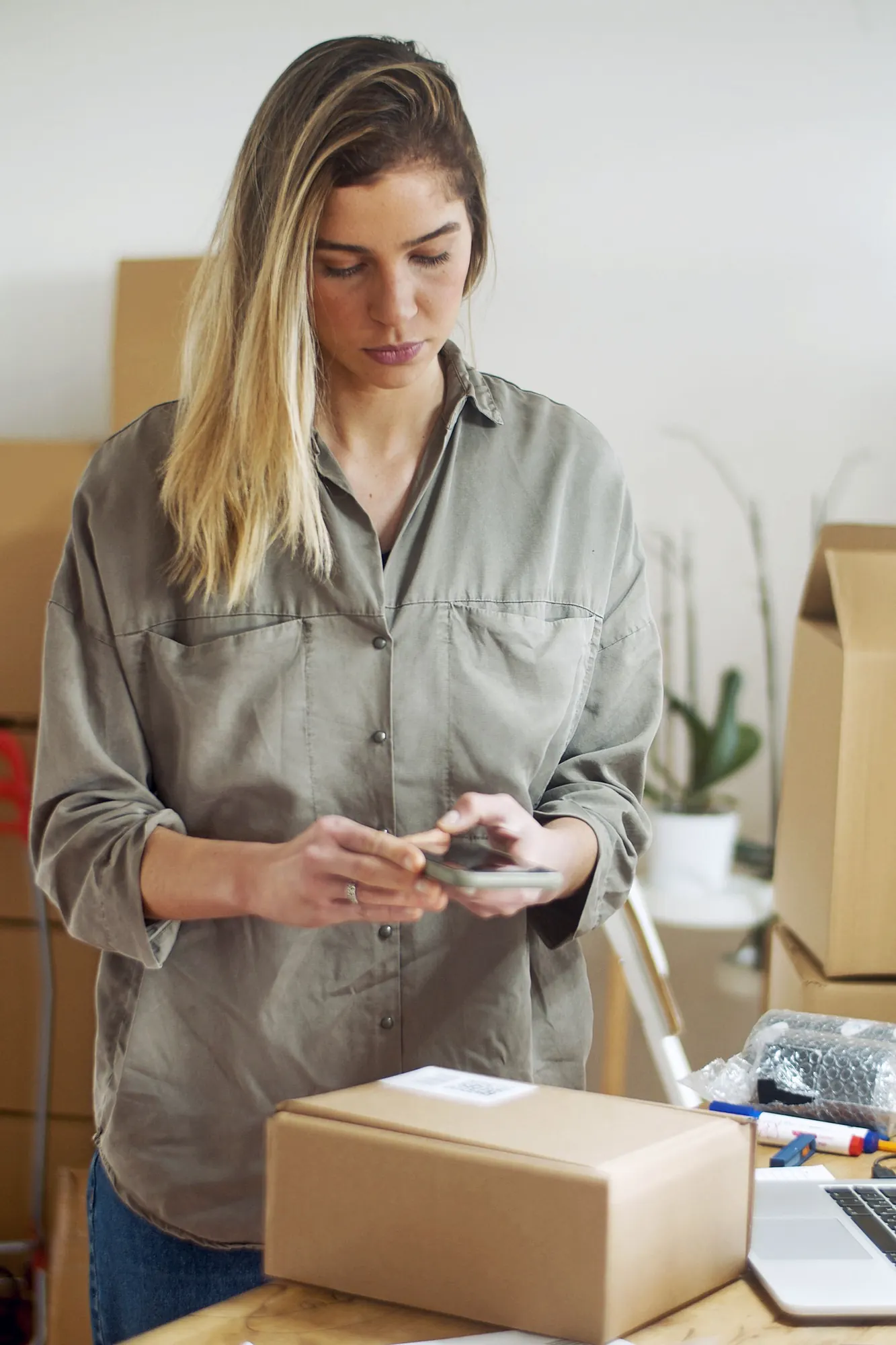 A small online seller checks an order on her phone over a packed shipping box in her home workspace