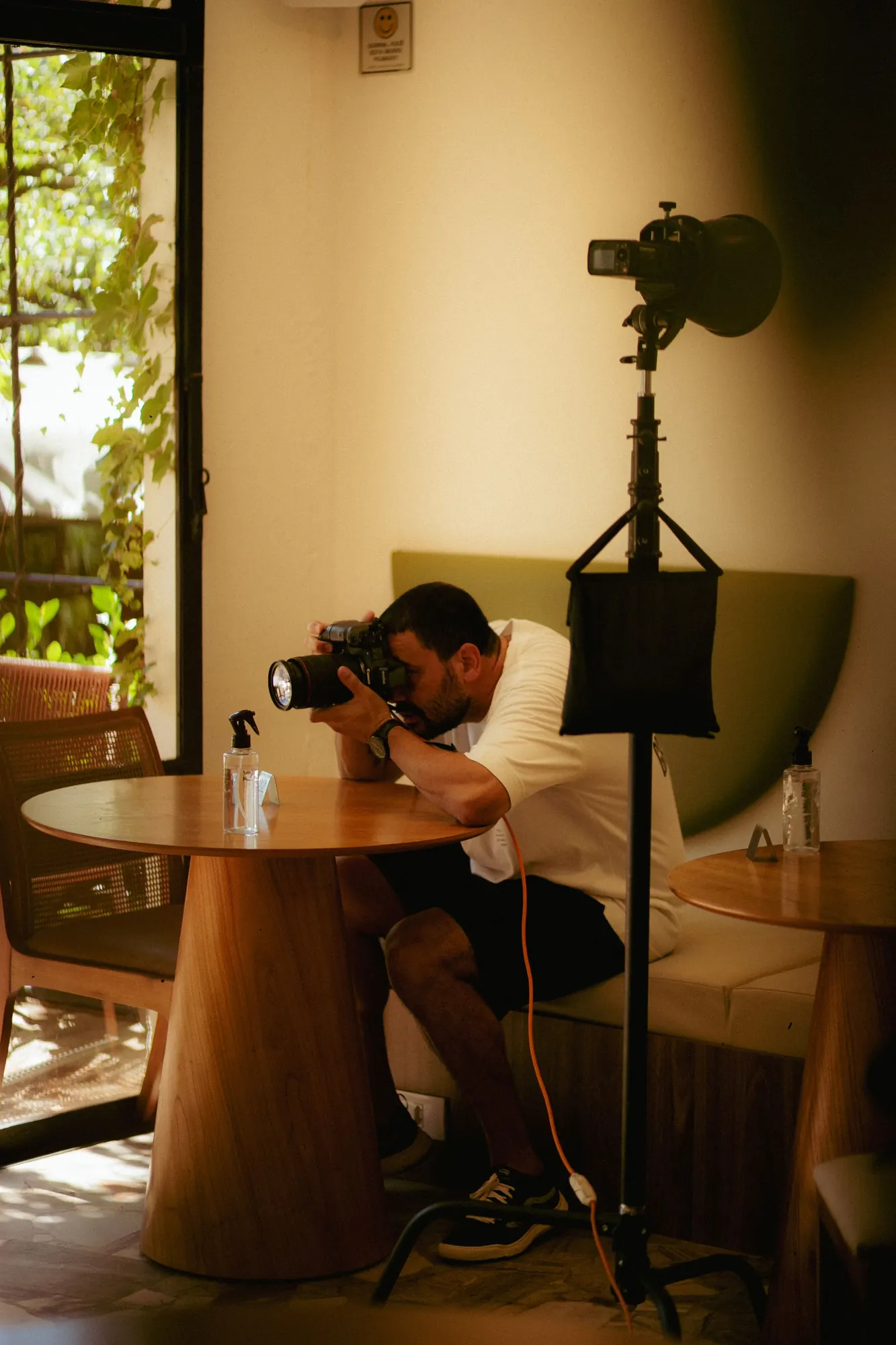A photographer crouches to shoot a cafe table set beside a light stand, in warm window light