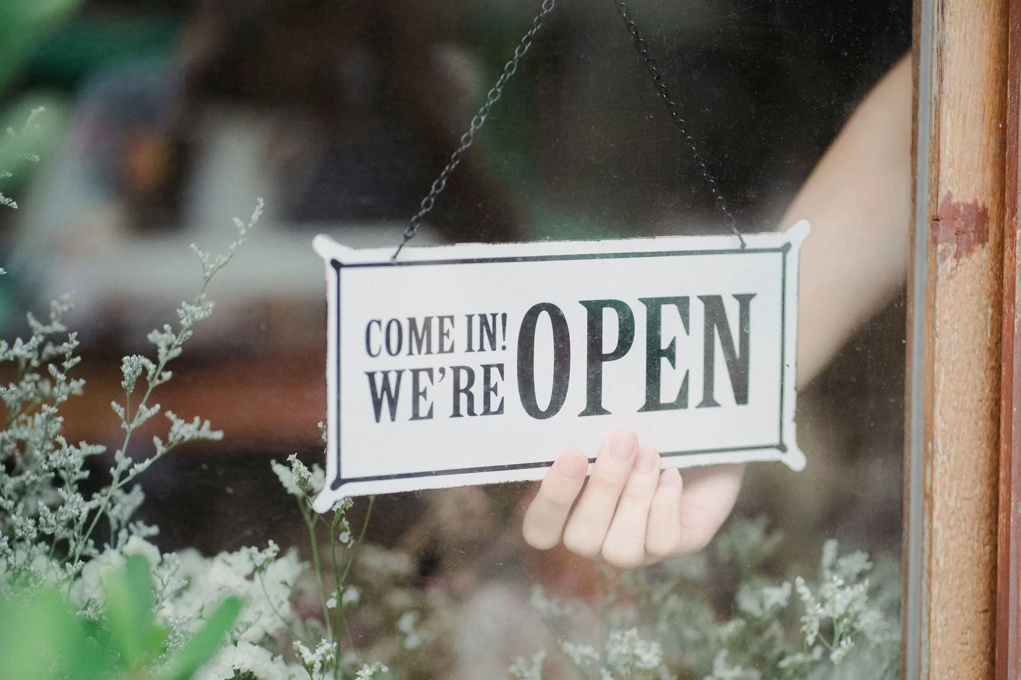 A hand hangs a "Come in! We're open" sign in a flower shop window behind dried flowers
