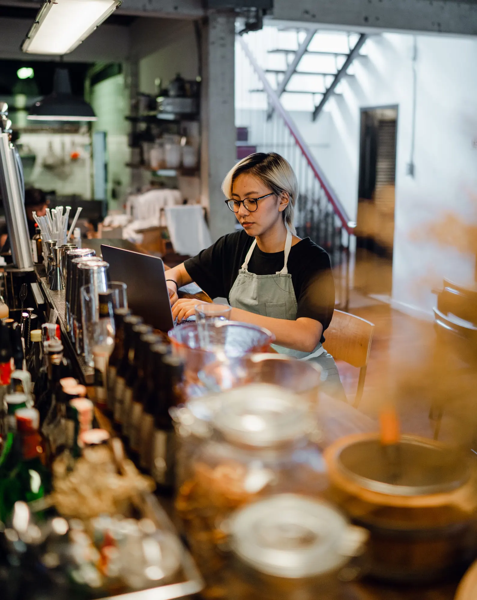A cafe-bar worker in an apron and glasses types on a laptop at her bar counter, with jars and bottles in the foreground