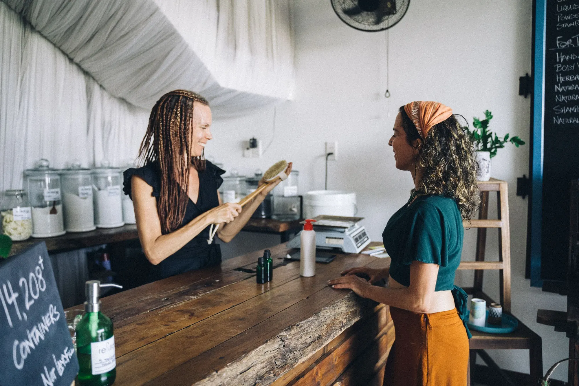 A shop owner shows a wooden brush to a customer across a worn wooden counter in a refill shop with glass jars