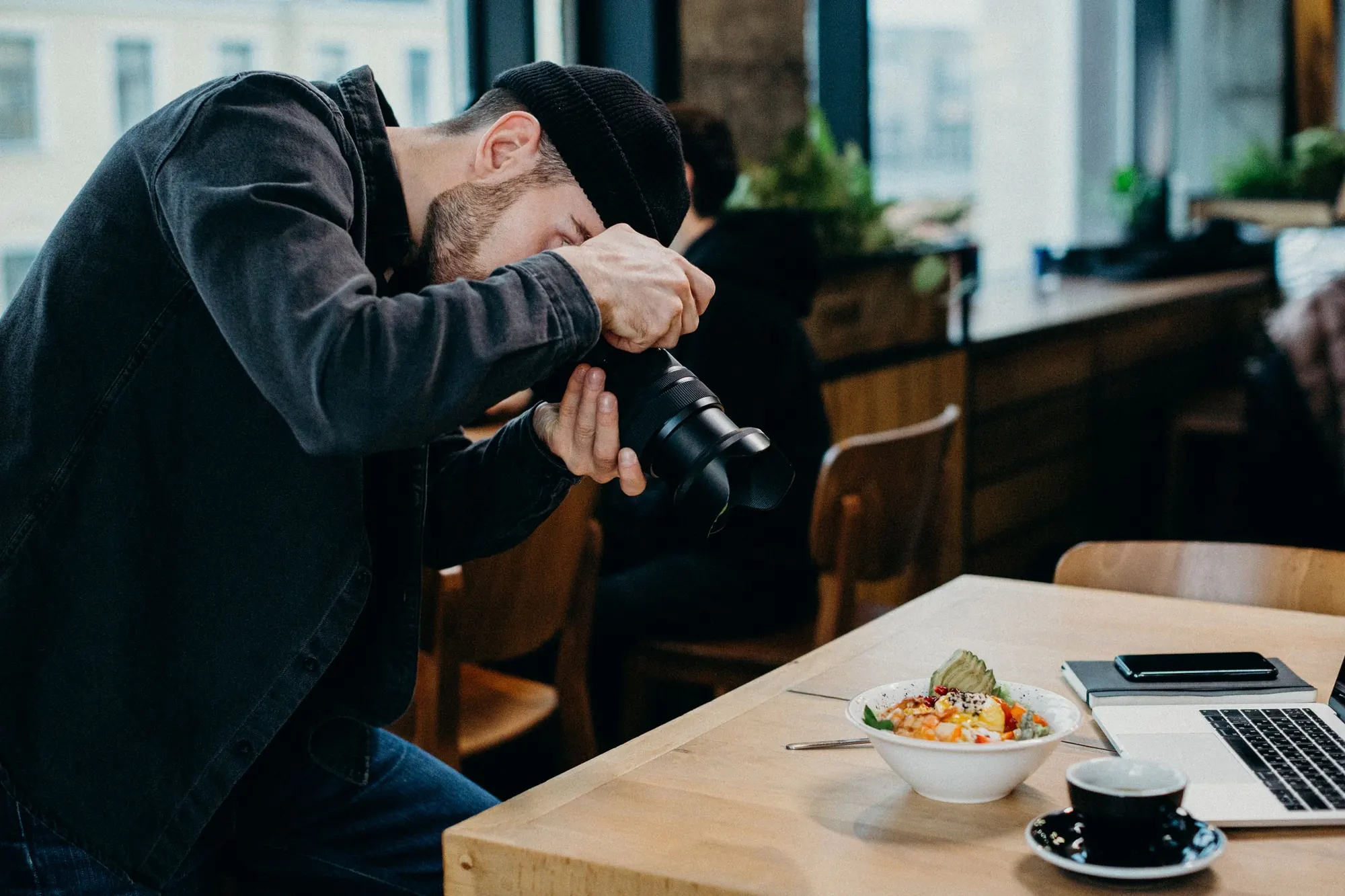A photographer bends over a cafe table to shoot a plated bowl, with a coffee cup and laptop beside it, by the window
