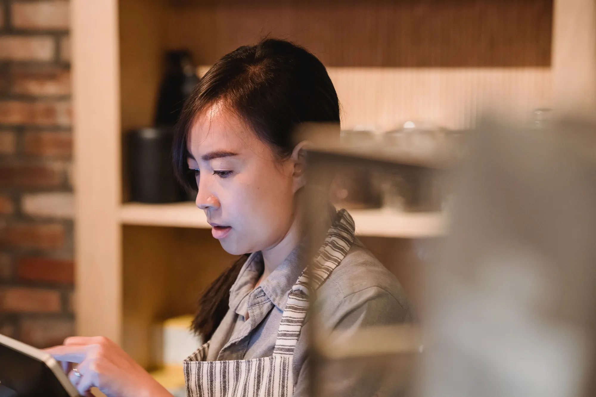 A cafe owner in a striped apron reviews a tablet behind the counter in warm light