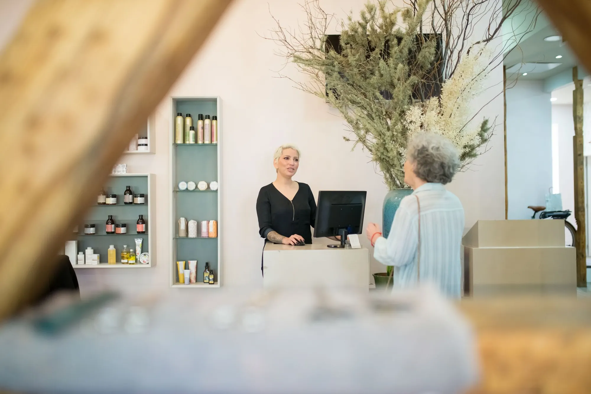 An older client chats with the receptionist at a salon's front desk, seen through a blurred foreground frame