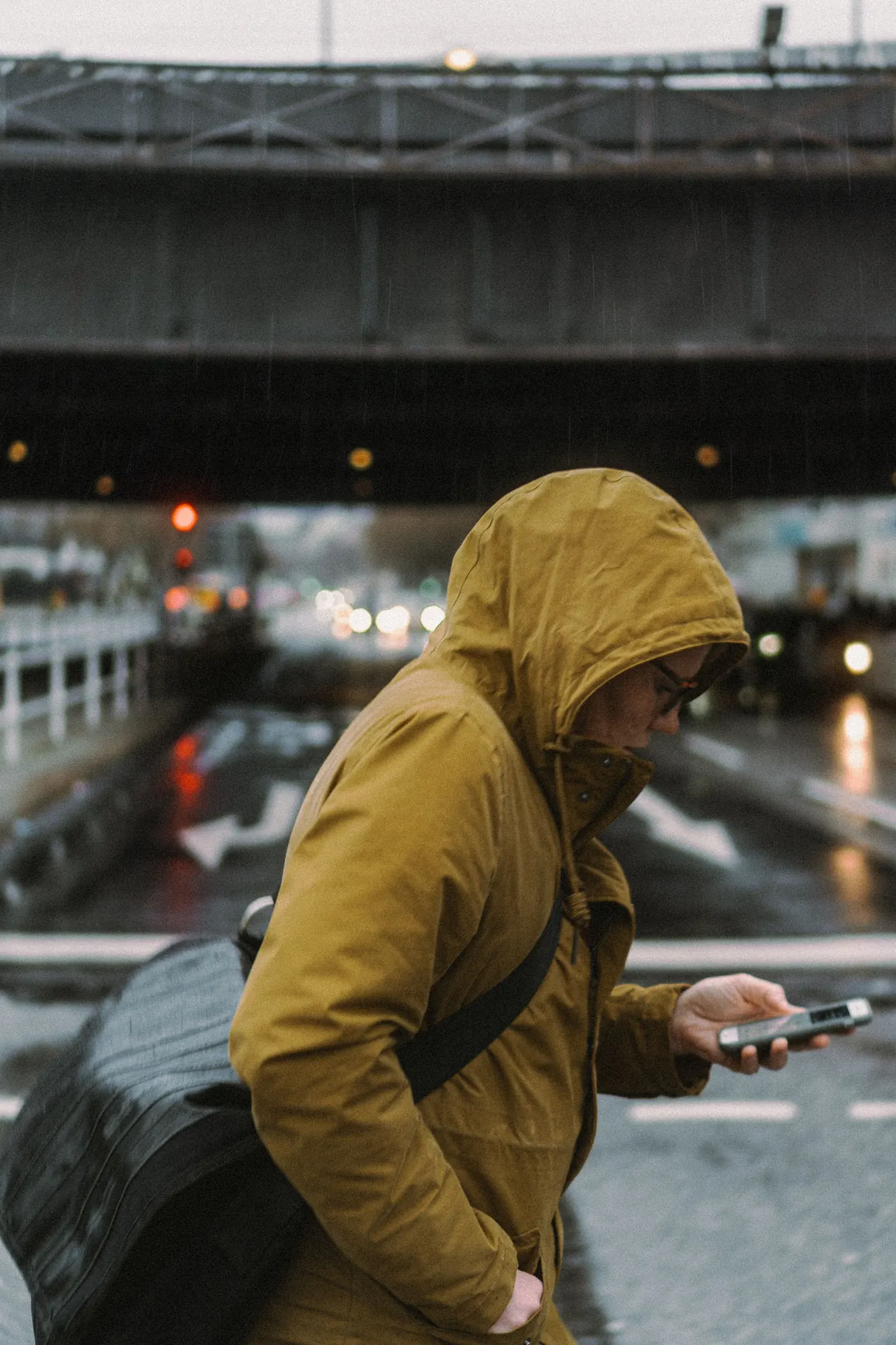 A person in a mustard hooded rain jacket checks their phone on a wet city street under an overpass